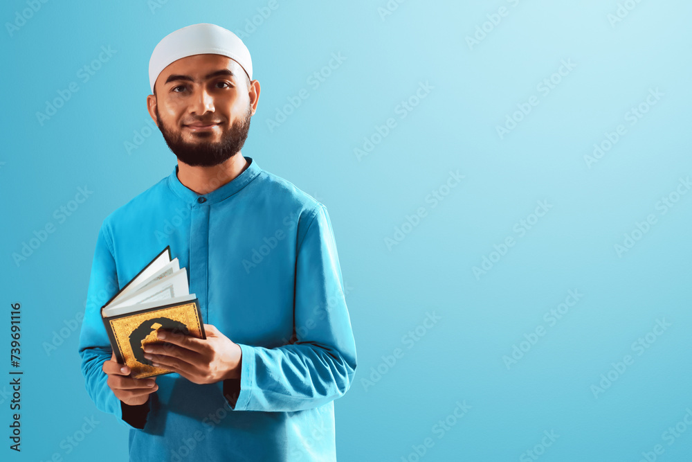 Portrait of handsome young asian muslim man with beard posing, holding holy book quran and smiling isolated on blue studio background Portrait of handsome young asian muslim man with beard posing, holding holy book quran and smiling isolated on blue studio background