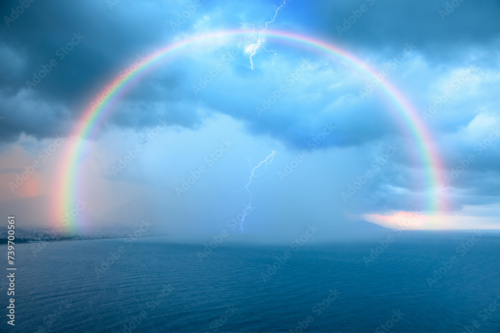 Stormy and rainy weather thunder and lightning strikes between clouds, rainbow in the background Stormy and rainy weather thunder and lightning strikes between clouds, rainbow in the background
