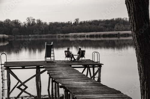 Couple sitting on a pier above the lake in sunset. Love concept. Black and white photo. About 30 years old. photo