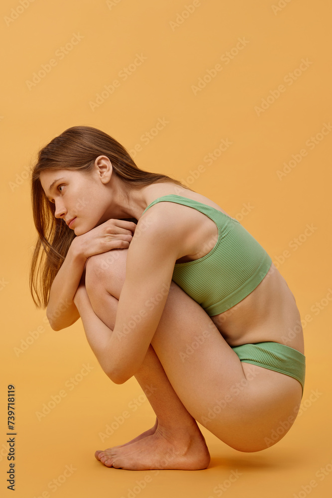 Vertical side view of young gen Z woman in pale green underwear posing for camera hugging her knees, warm apricot color background