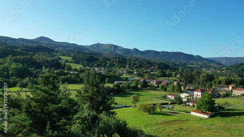 drone flight in ascent looking at the treetops in the Basque-Cantabra area over a rural town with farmland and lush oak forests and mountains on an afternoon with blue sky in summer and green grass