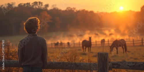 Portrait of a young man 17-year-old in the country standing with his back to us near a low fence, Behind the fence, horses roamed under the soft sunlight photo