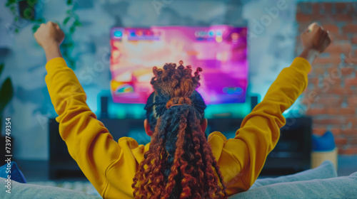 Young black African American woman enjoy a victory playing video games on TV screen raising her arms in the air on her home couch photo
