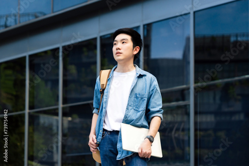 Portrait of handsome Asian student holding computer laptop and backpack. A young man standing outdoor happy smiling