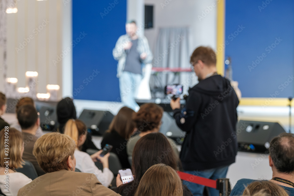 Obraz premium Defocused image of a speaker at event with audience watching intently, capturing the atmosphere of live presentations.