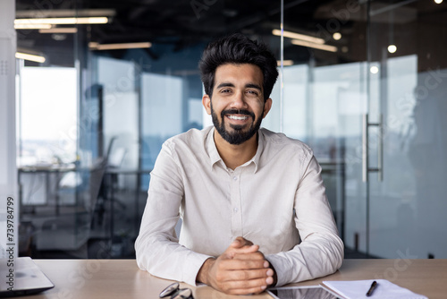 Portrait of a young Indian man in a shirt sitting smiling at a desk in the office and looking confidently at the camera photo
