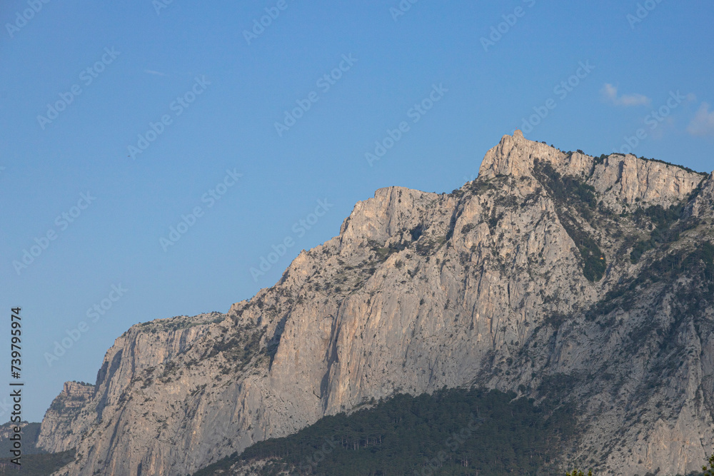 The peak, high gray cliffs against the blue sky, the forest at the foot of the mountains. Mountain scenery.