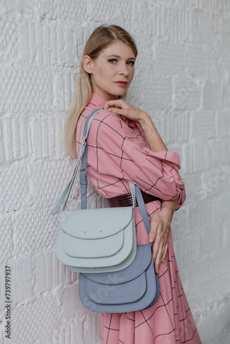 A beautiful slender girl in a pink dress poses against a white wall, she holds two handbags in her hands