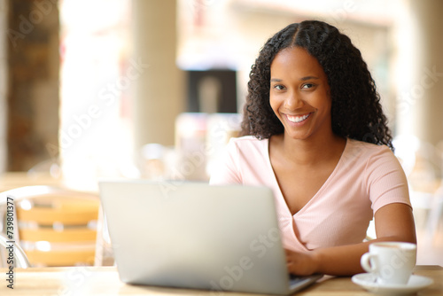 Happy black woman looking at you using laptop in a restaurant