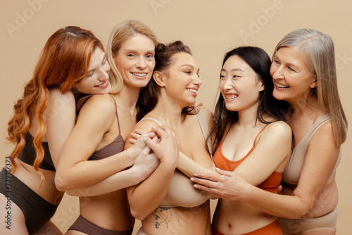 Group of happy, beautiful, multiracial women wearing sexy lingerie, hugging, looking at camera, posing isolated on beige background. Support concept, international women's day