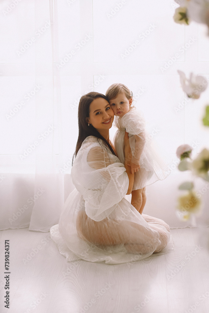 Studio portrait of smiling mother in white dress sitting near window and holding her baby daughter in her arms Studio portrait of smiling mother in white dress sitting near window and holding her baby daughter in her arms