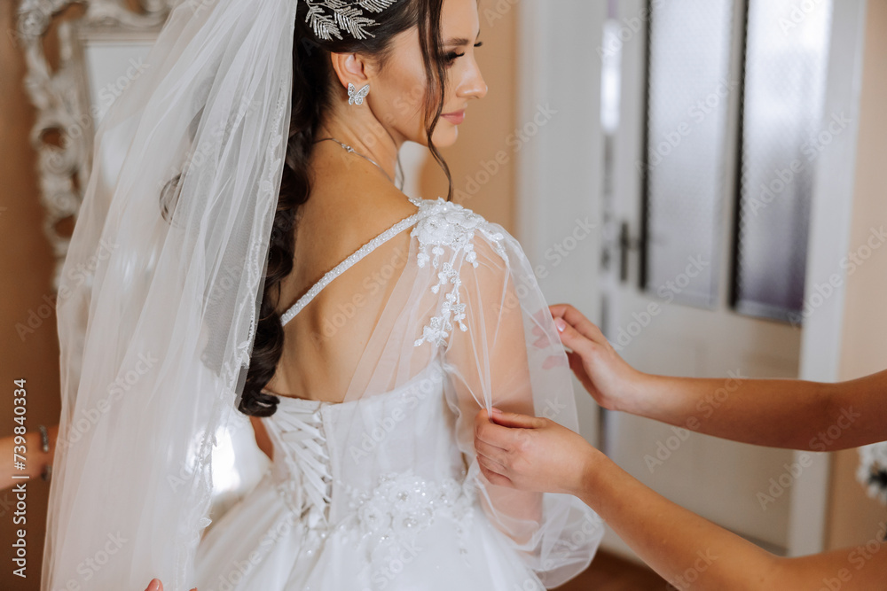 The bride's friend helps her get dressed in a white wedding dress in the morning. wedding morning, blurred focus. A friend of the bride helps to tie her wedding dress. The bride's friend helps her get dressed in a white wedding dress in the morning. wedding morning, blurred focus. A friend of the bride helps to tie her wedding dress.