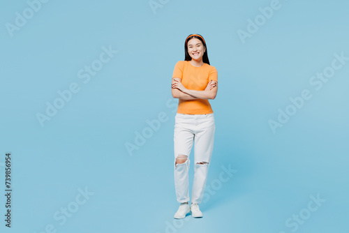 Full body smiling happy young woman of Asian ethnicity she wear orange t-shirt casual clothes hold hands crossed folded isolated on pastel light blue cyan background studio portrait Lifestyle concept.