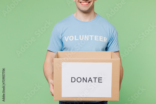Cropped close up young man wear blue t-shirt white title volunteer hold cardboard donation box isolated on plain pastel green background Voluntary free team work assistance help charity grace concept photo