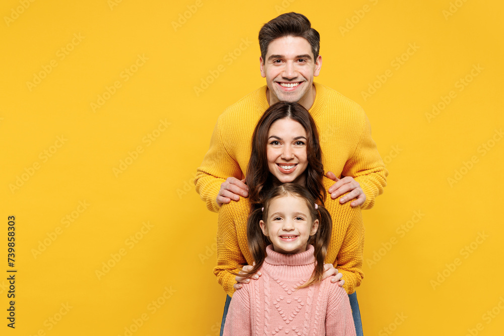 Young happy smiling cheerful parents mom dad with child kid girl 7-8 years old wear pink knitted sweater casual clothes stand behind each other isolated on plain yellow background. Family day concept. Young happy smiling cheerful parents mom dad with child kid girl 7-8 years old wear pink knitted sweater casual clothes stand behind each other isolated on plain yellow background. Family day concept.