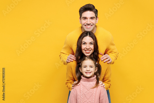Young happy smiling cheerful parents mom dad with child kid girl 7-8 years old wear pink knitted sweater casual clothes stand behind each other isolated on plain yellow background. Family day concept. photo