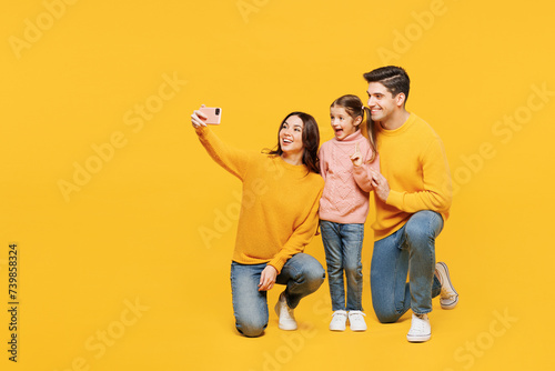 Full body young happy parents mom dad with child kid girl 7-8 years old wear pink sweater casual clothes doing selfie shot on mobile cell phone isolated on plain yellow background. Family day concept. photo