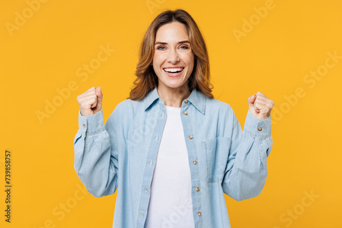 Young happy cool woman she wear blue shirt white t-shirt casual clothes doing winner gesture celebrate clenching fists say yes isolated on plain yellow background studio portrait. Lifestyle concept.