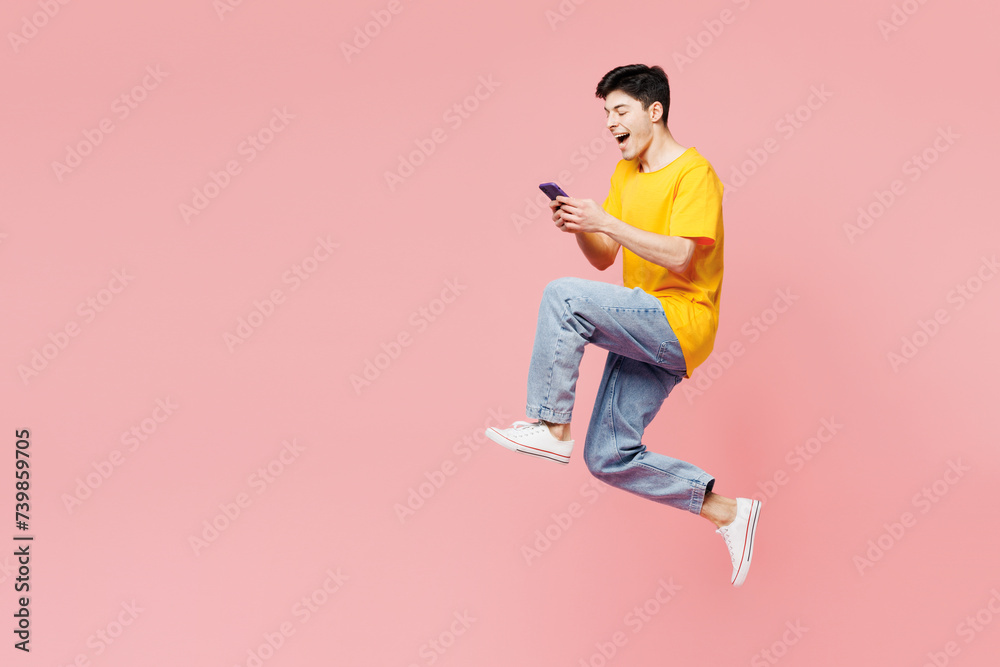 Full body side profile view excited young man wear yellow t-shirt casual clothes jump high hold use mobile cell phone isolated on plain pastel light pink background studio portrait. Lifestyle concept Full body side profile view excited young man wear yellow t-shirt casual clothes jump high hold use mobile cell phone isolated on plain pastel light pink background studio portrait. Lifestyle concept