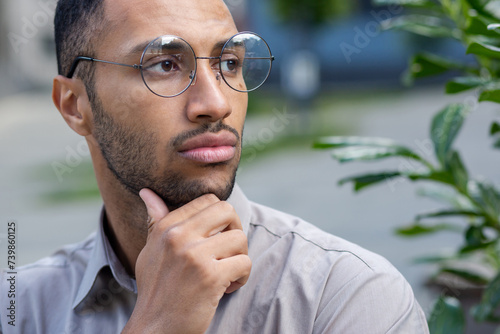 A close-up photo of a young Hispanic man standing outside the city wearing glasses, rubbing his beard with his hand, looking seriously and thoughtfully to the side.