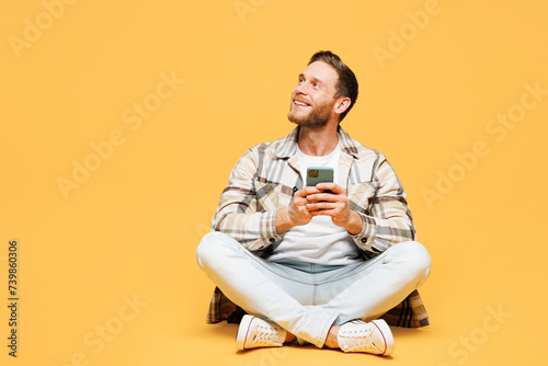 Full body young Caucasian man he wears brown shirt casual clothes sitting hold in hand use mobile cell phone look aside isolated on plain yellow orange background studio portrait. Lifestyle concept. © ViDi Studio