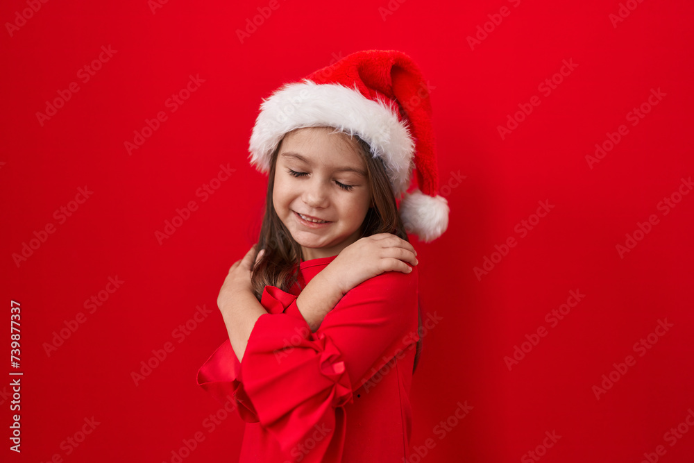 Adorable hispanic girl, all smiles, hugging herself in self-love, radiating happiness over an isolated red wall. donned in a christmas hat, exuding confidence and positive vibes. Adorable hispanic girl, all smiles, hugging herself in self-love, radiating happiness over an isolated red wall. donned in a christmas hat, exuding confidence and positive vibes.
