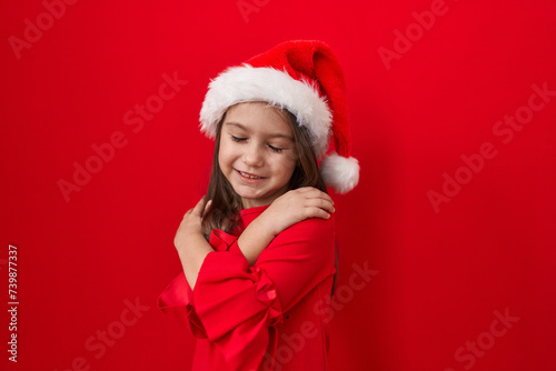 Adorable hispanic girl  all smiles  hugging herself in self-love  radiating happiness over an isolated red wall. donned in a christmas hat  exuding confidence and positive vibes.