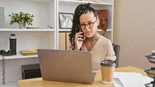 Successful beautiful young hispanic woman worker engrossed in a serious business talk over smartphone, working on laptop amidst the hustle and bustle of indoor office environment