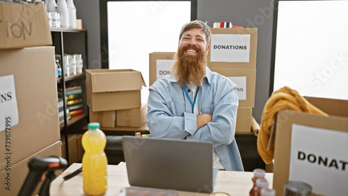 Confident young redhead man, arms crossed, volunteering at charity center, working away on laptop, sitting at table with heartwarming smile