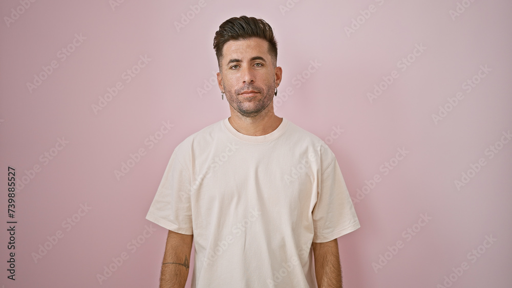 Cool young hispanic man, a relaxed yet concentrated expression marking his handsome face. he's standing casually to the side, isolated over a pink background, looking seriously into the camera Cool young hispanic man, a relaxed yet concentrated expression marking his handsome face. he's standing casually to the side, isolated over a pink background, looking seriously into the camera