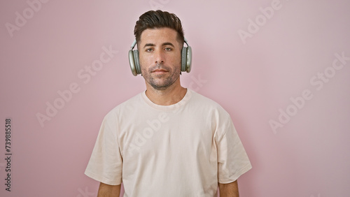 Cool, young hispanic man, looking serious, totally immersed, listening to a song over headphones. portrait of attractive guy against isolated pink wall, techno gadget in hand. © Krakenimages.com