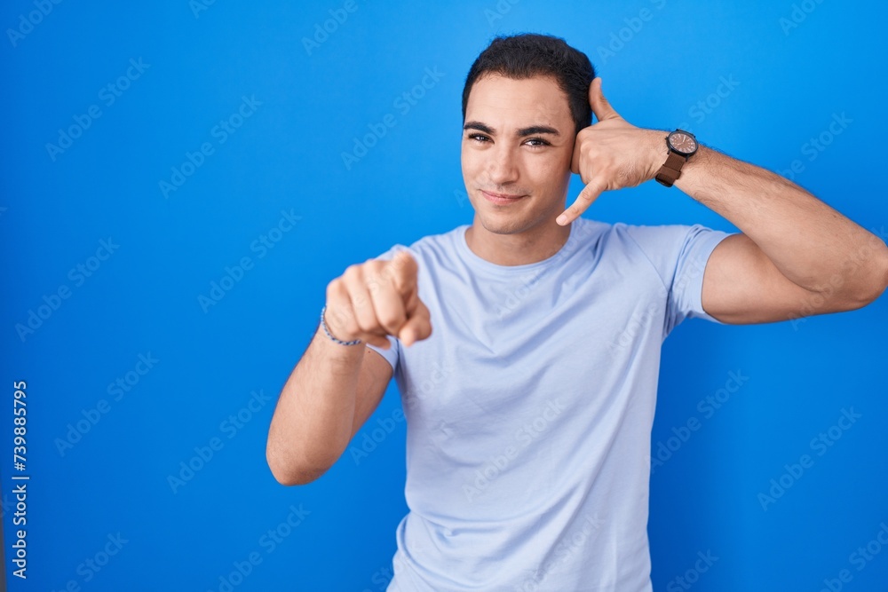 Young hispanic man standing over blue background smiling doing talking on the telephone gesture and pointing to you. call me. Young hispanic man standing over blue background smiling doing talking on the telephone gesture and pointing to you. call me.