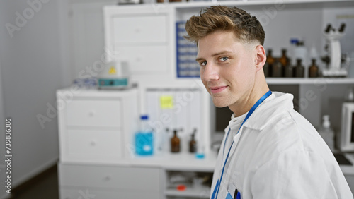 Smiling confident caucasian young man  a handsome scientist  sitting in his lab enjoying the research work. indoors  surrounded by test tubes  a microscope - totally in his element 