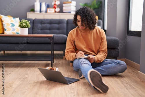 Young latin man using laptop writing on notebook at home