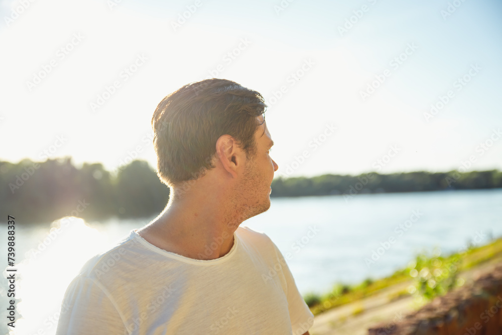 young adult man looking away on city beach at sunset on river. man looks thoughtfully into distance, watching
