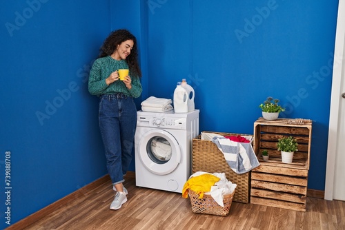 Young beautiful hispanic woman drinking coffee waiting for washing machine at laundry room