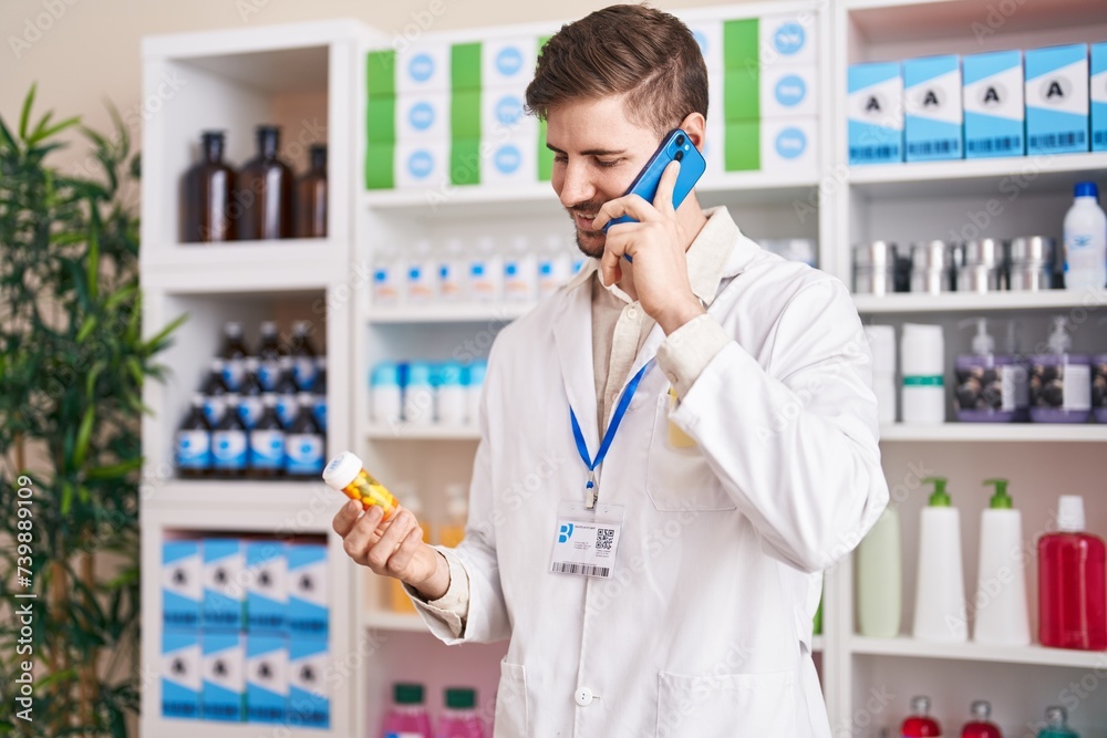 Young caucasian man pharmacist holding pills bottle talking on smartphone at pharmacy