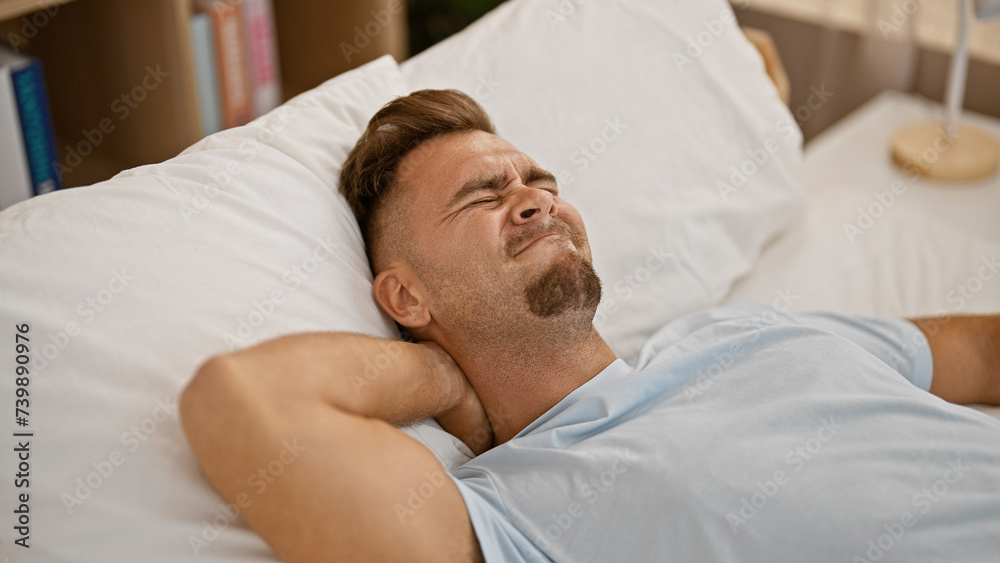 Obraz premium Young hispanic man with a beard showing discomfort in a bedroom setting, lying on white bedding, expressing pain.