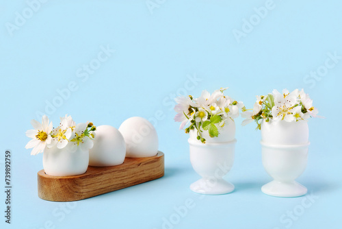 White eggs in wooden and white stands with spring pear and strawberry flowers on a blue background. Happy Easter