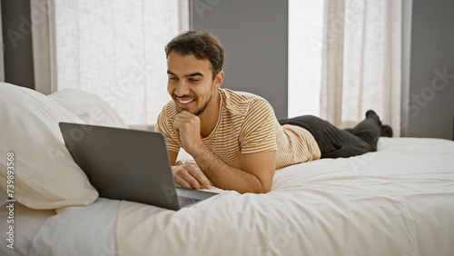 A smiling young man lies on a bed with a laptop in a bright bedroom, indicating comfort and leisure at home.