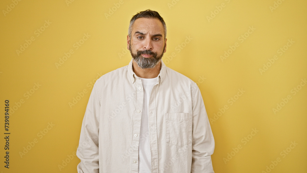 A mature hispanic man with grey beard stands confidently against an isolated yellow background.