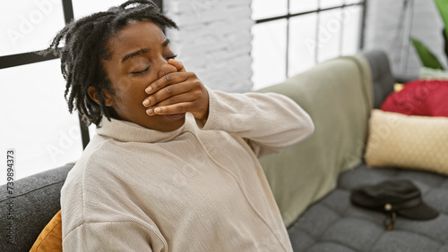 A young woman yawning, seated on a sofa indoors, exuding a relaxed, homey atmosphere. © Krakenimages.com