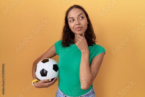 Young hispanic woman holding ball looking confident at the camera smiling with crossed arms and hand raised on chin. thinking positive. © Krakenimages.com