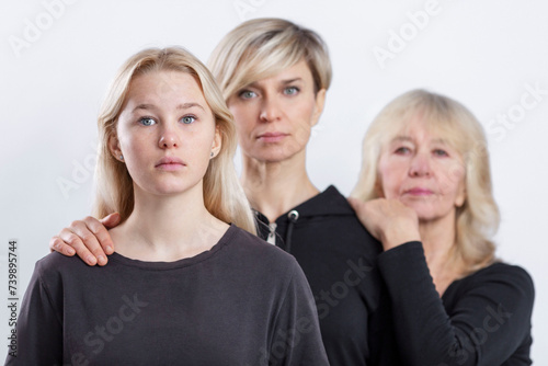 Portrait of three generations of blonde women in a family. Serious mother, grandmother and granddaughter in black sweaters stand behind each other. Selective focus. White background.