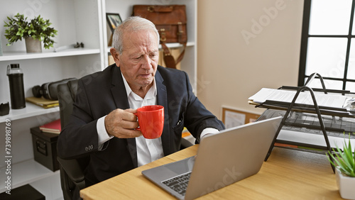 Elegant senior man, focused on success, working online with laptop at office desk, drinking morning coffee in a relaxed but serious way.