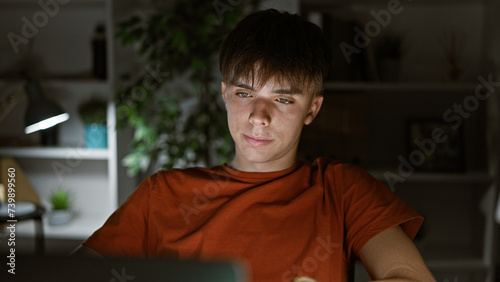 A focused young caucasian male teen works on his laptop in a dimly lit room at night, exemplifying contemporary indoor lifestyle.