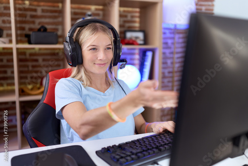 Young caucasian woman playing video games wearing headphones smiling cheerful offering palm hand giving assistance and acceptance.