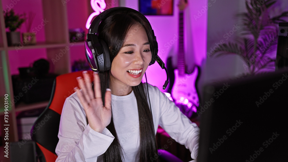 Smiling asian woman with headphones in neon-lit gaming room waving at computer screen
