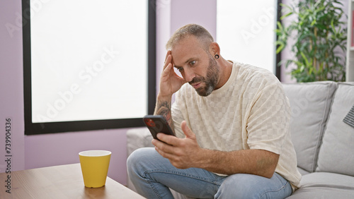 Surprised young man, sitting relaxed on sofa at home, drinking morning espresso from a mug, looking at his smartphone screen with amazed expression photo