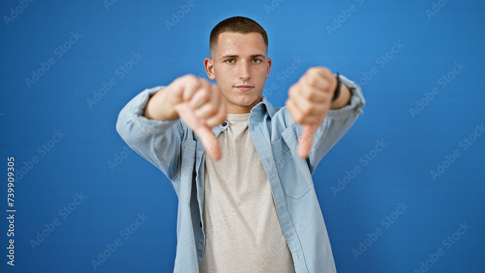 Hispanic man giving thumbs down gesture against a solid blue background, expressing disapproval